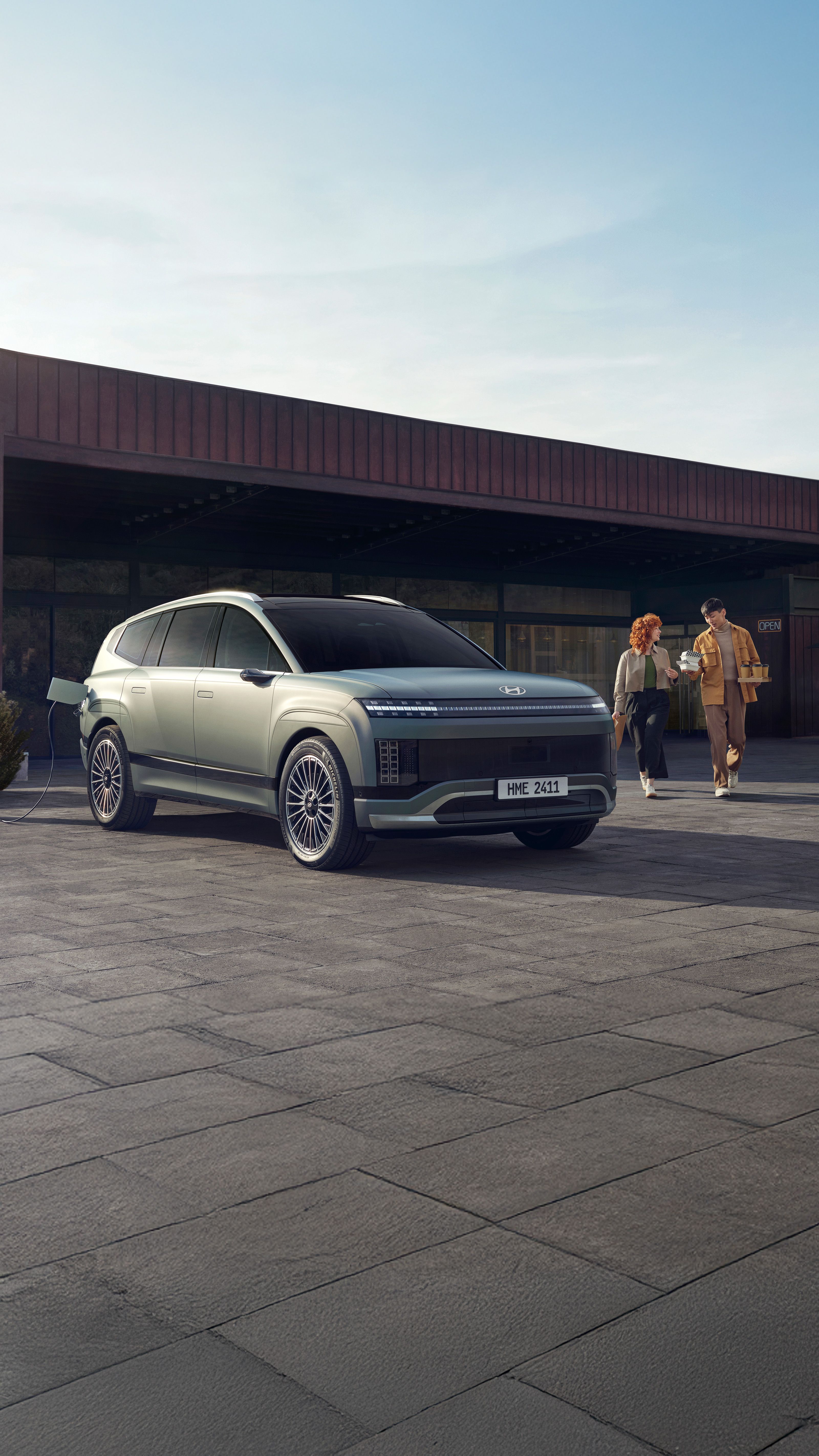 Et ungt par kommer gående med kaffe bak en IONIQ 9 som står og lader. Foto.